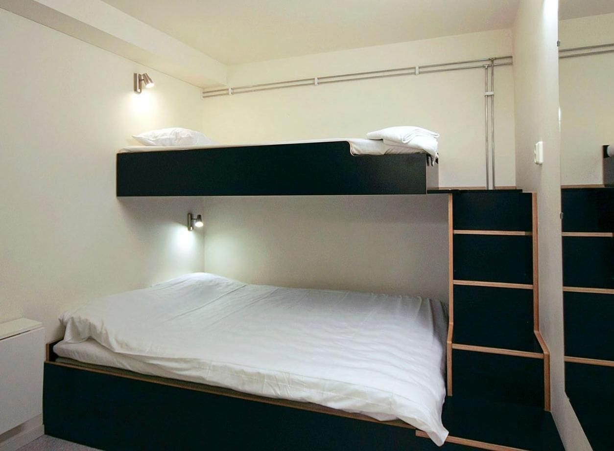A bunk bed with a white mattress and a black headboard. There is a small bedside table and a wall-mounted light fixture.