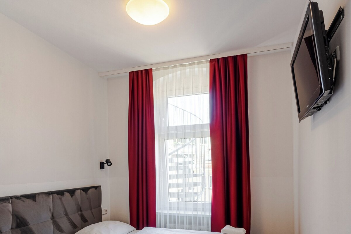 A hotel room with a double bed, a flat-screen TV mounted on the wall, and a window with red curtains.