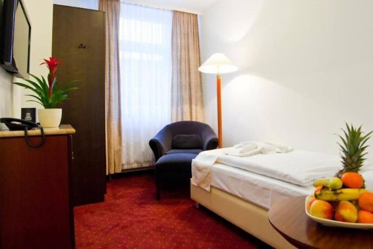 A hotel room with a bed, a desk with a telephone, a chair, a bedside table with fruit, and a lamp.