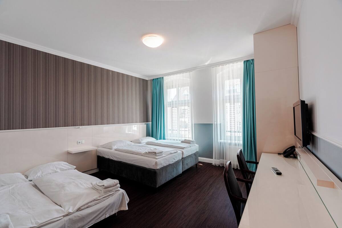 A hotel room with two single beds, a desk with a chair, a TV, and a window with curtains.