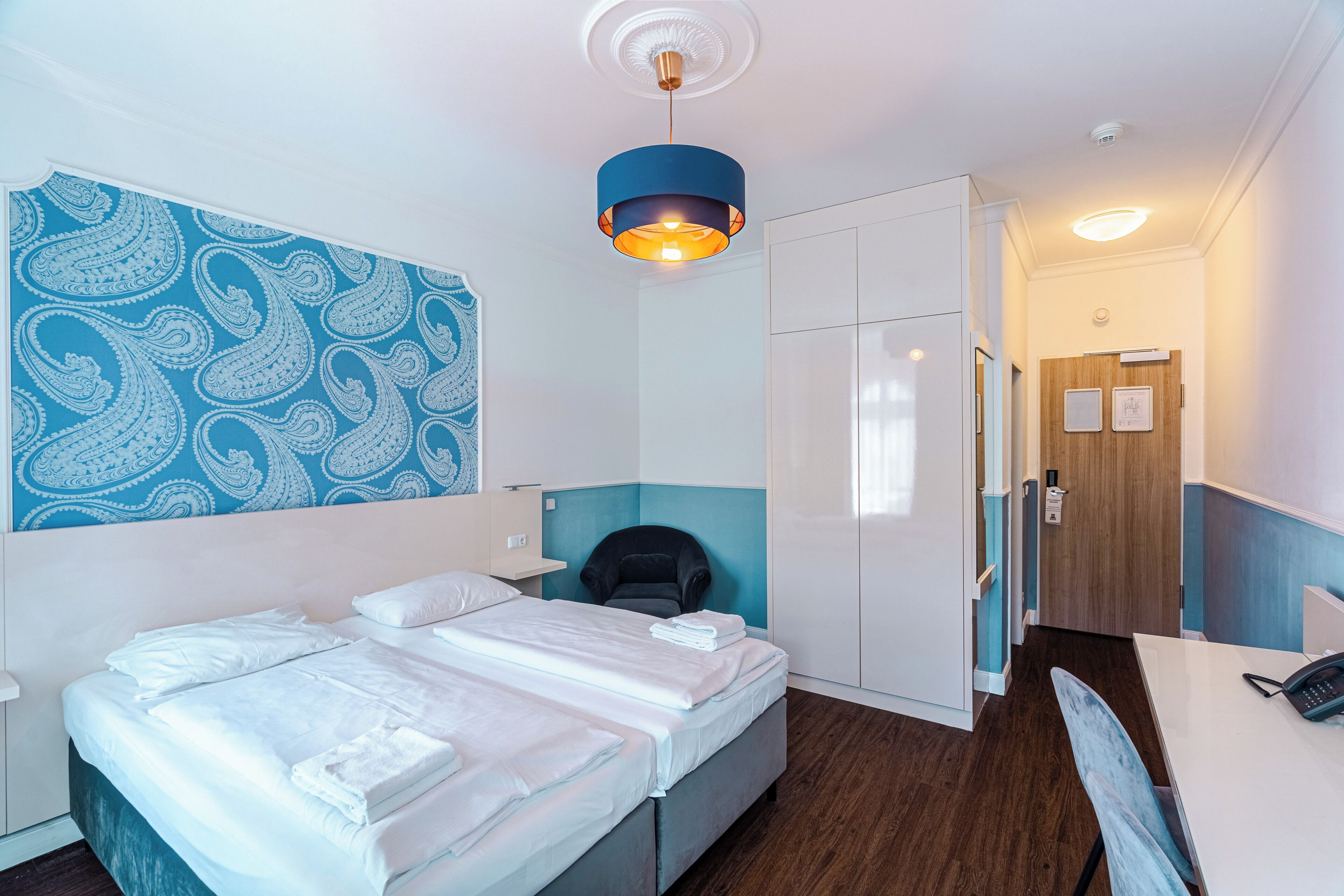 Een hotelkamer met een bed, een bureau, een stoel, een kledingkast en een muur met een paisley dessin.