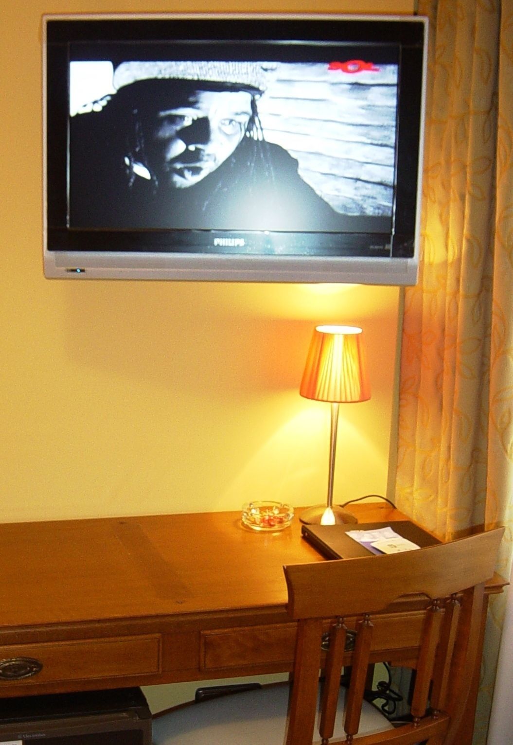 Un televisor montado en la pared, un escritorio de madera con una lámpara y una silla.