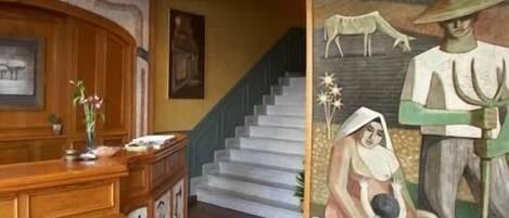 Un vestíbulo con un mostrador de recepción de madera, una escalera y un mural grande que representa una escena campestre.