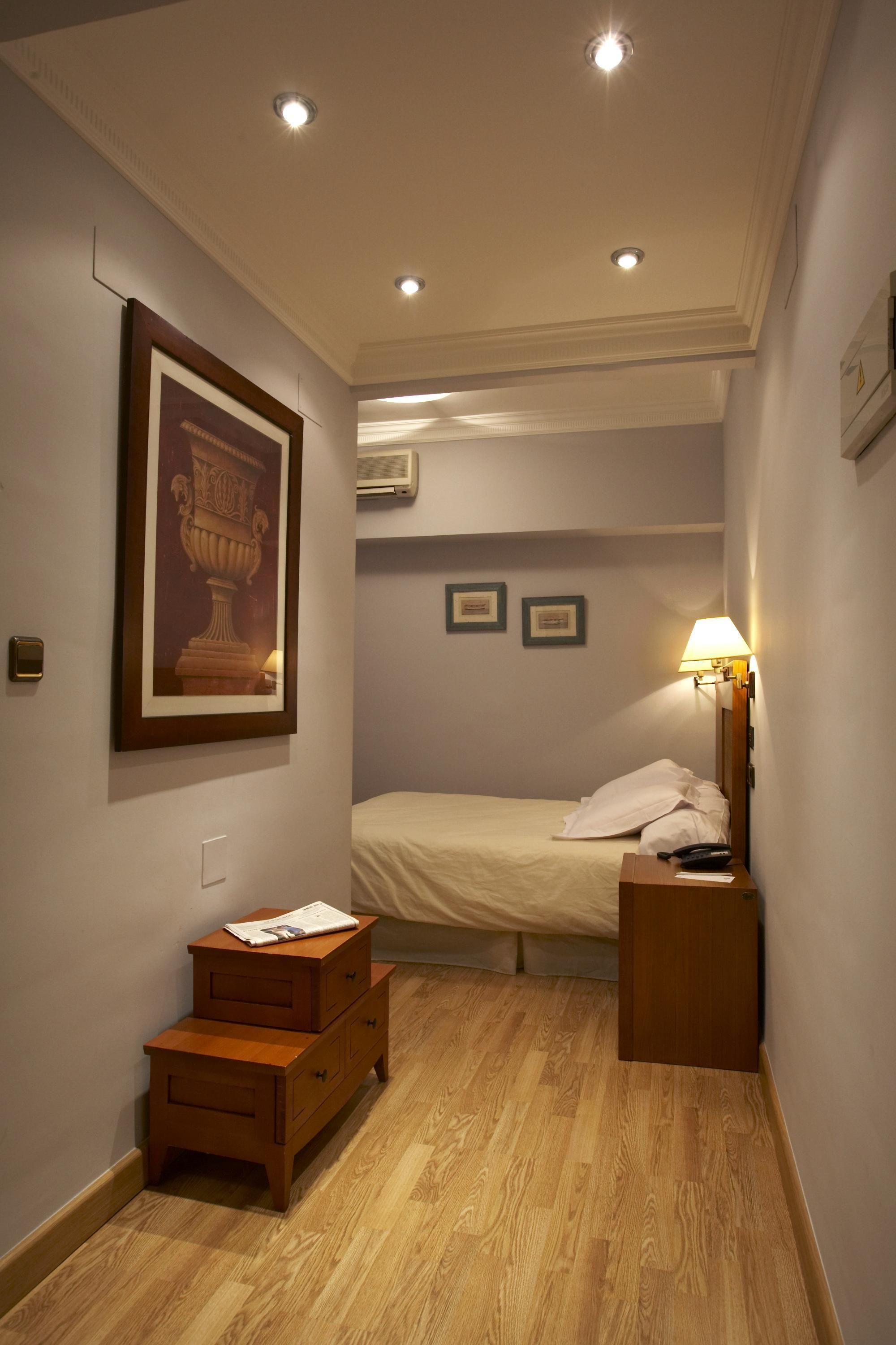 Un dormitorio con cama, mesita de noche, cómoda de madera, un cuadro en la pared y luces empotradas en el techo.