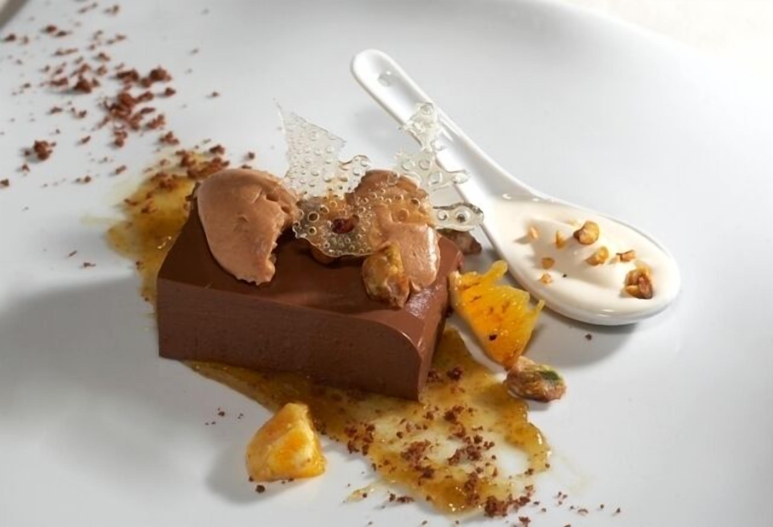 A dessert with a chocolate base, caramel, and a white sauce, accompanied by a spoon and scattered nuts.
