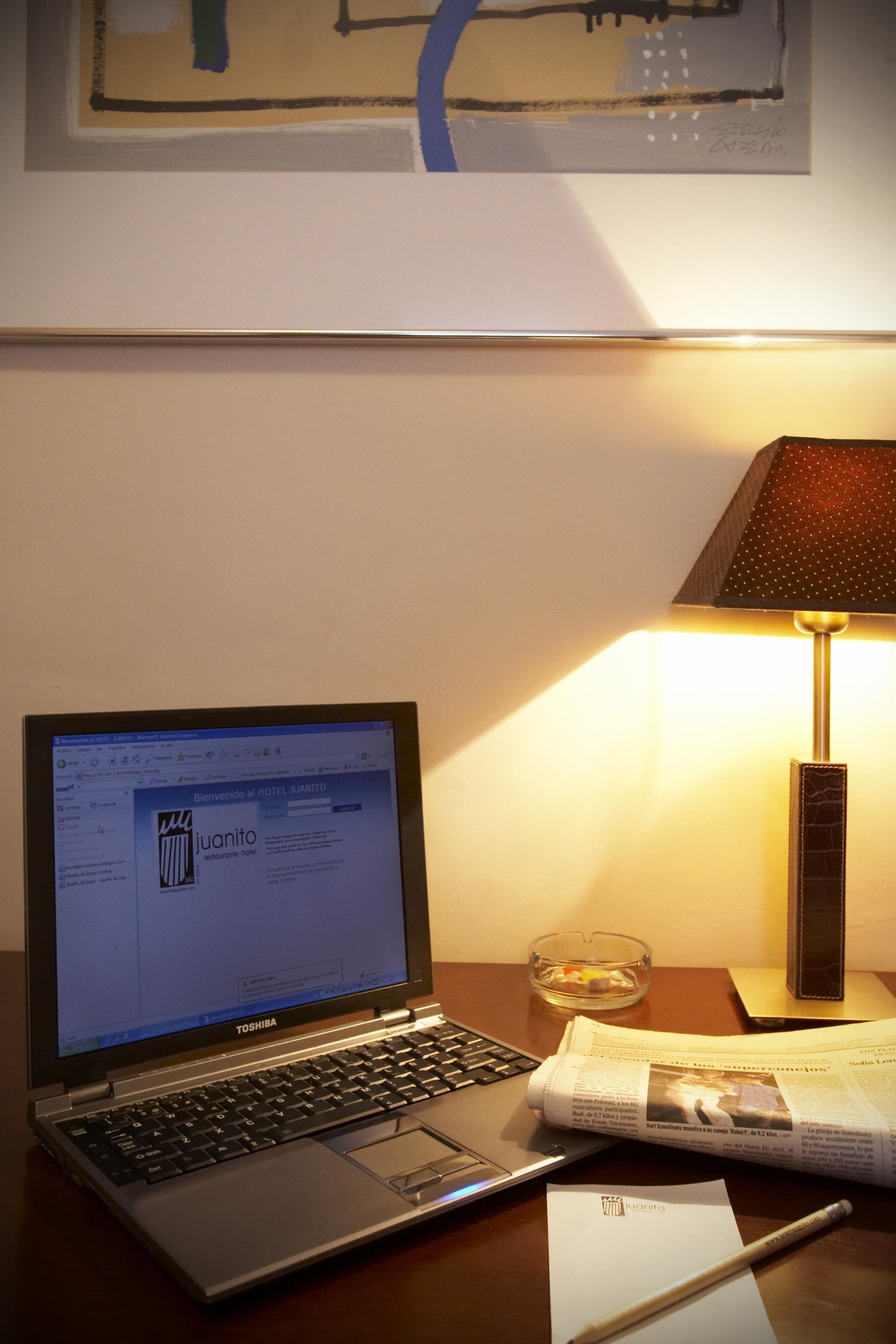 Um laptop Toshiba sobre uma mesa, com um jornal, uma caneta e um cinzeiro de vidro.