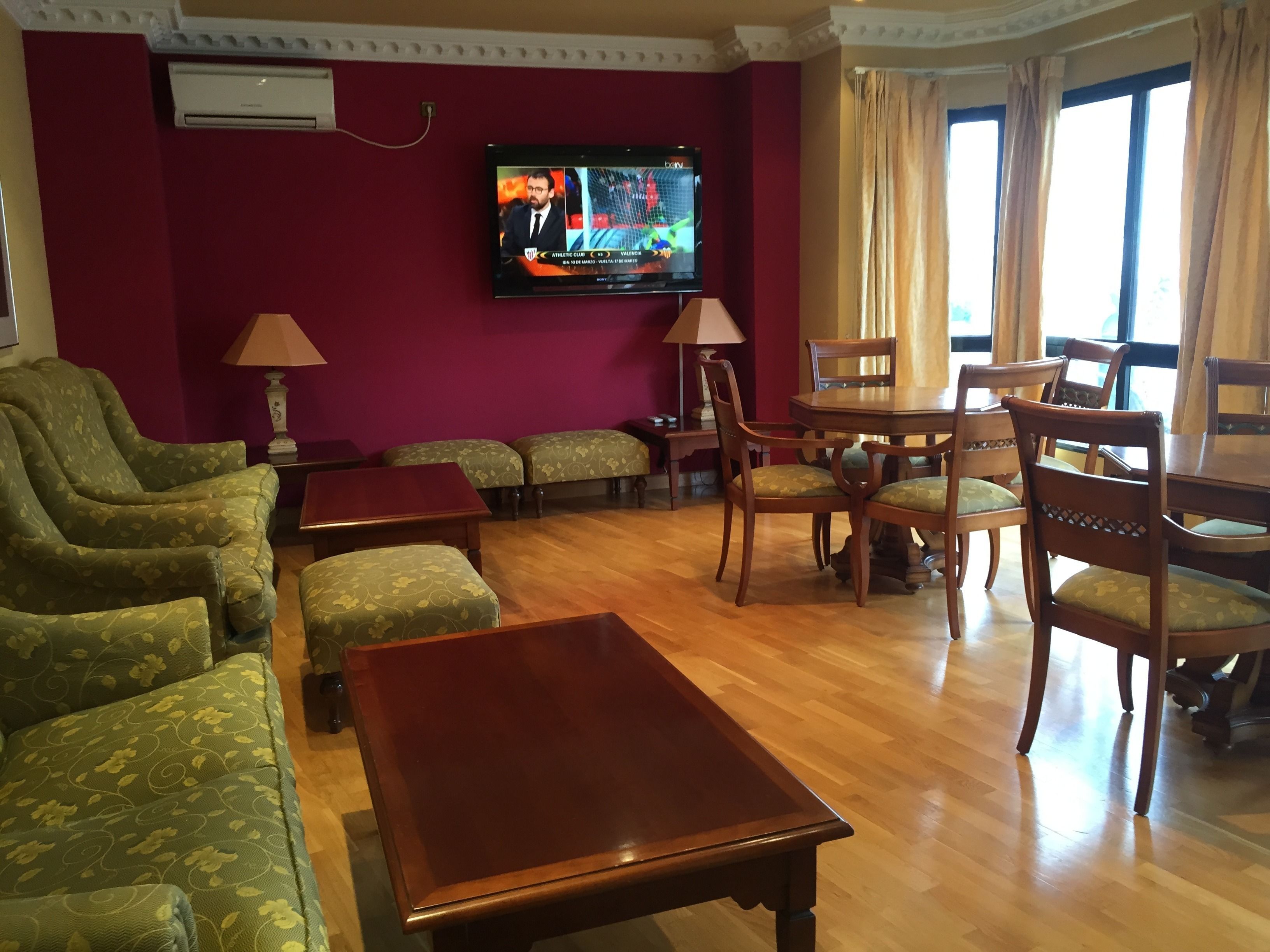A hotel room with a TV, green patterned sofas, wooden chairs, a wooden table, and a window with curtains.