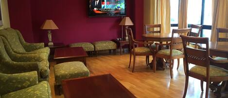 A hotel room with a TV, green patterned sofas, wooden chairs, a wooden table, and a window with curtains.