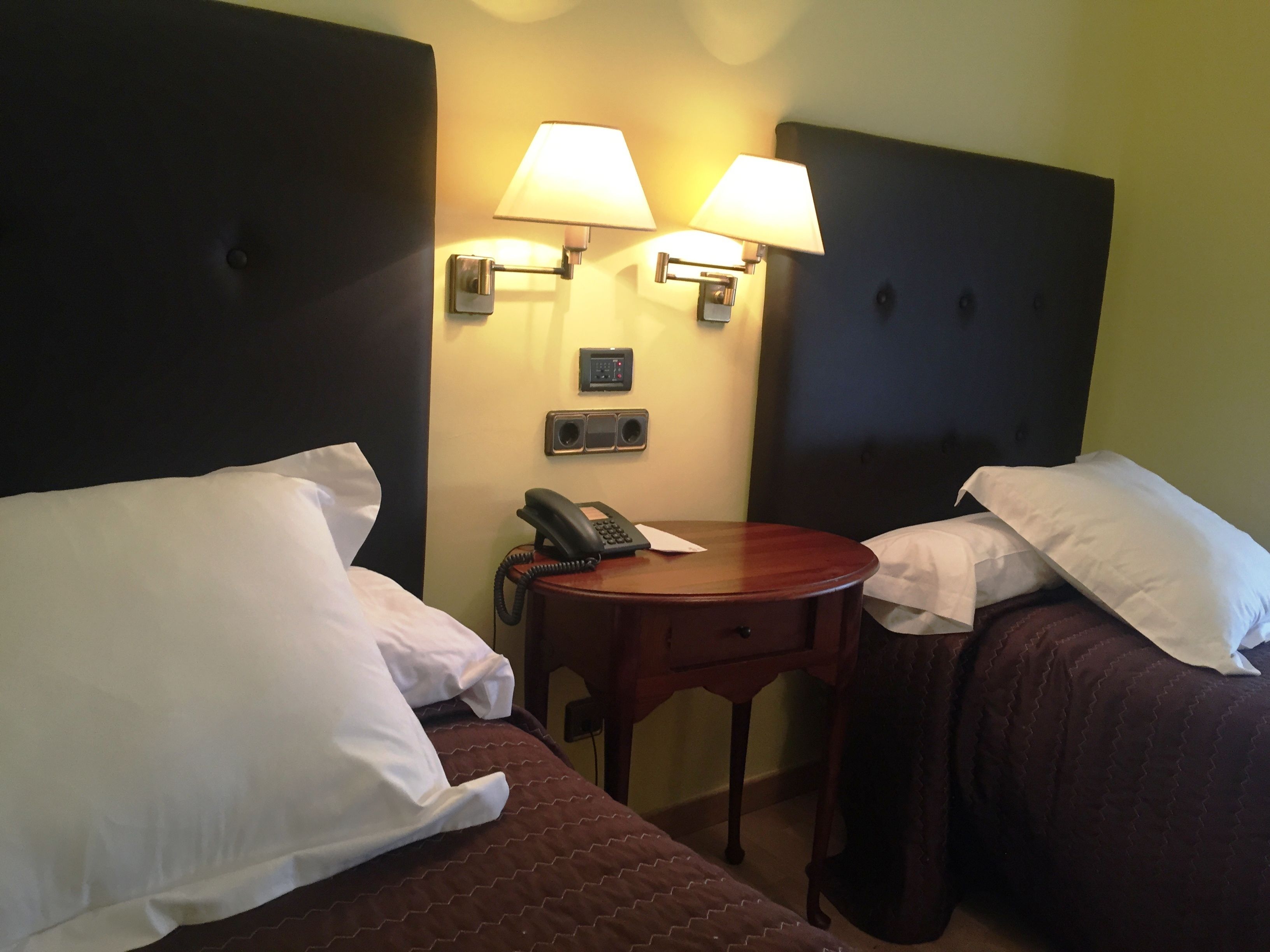 Habitación de hotel con dos camas, cada una con almohadas blancas y una colcha acolchada marrón. Hay dos lámparas de pared, un teléfono sobre una mesa de madera y un reloj pequeño en la pared.
