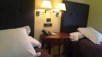 Habitación de hotel con dos camas, cada una con almohadas blancas y una colcha acolchada marrón. Hay dos lámparas de pared, un teléfono sobre una mesa de madera y un reloj pequeño en la pared.