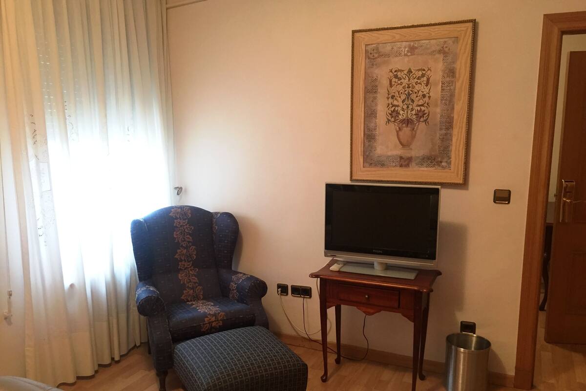 A room with a blue armchair, a flat-screen TV on a wooden stand, a framed artwork on the wall, and a door to the right.