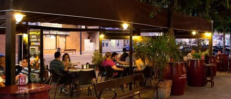 An outdoor café with tables and benches under a canopy, surrounded by potted plants and a street view.