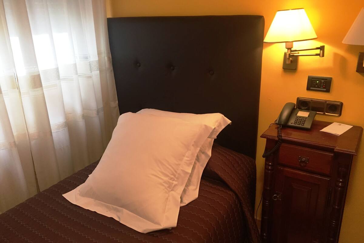 A hotel room with a bed, a bedside table with a lamp, a phone, and a wall-mounted light switch.