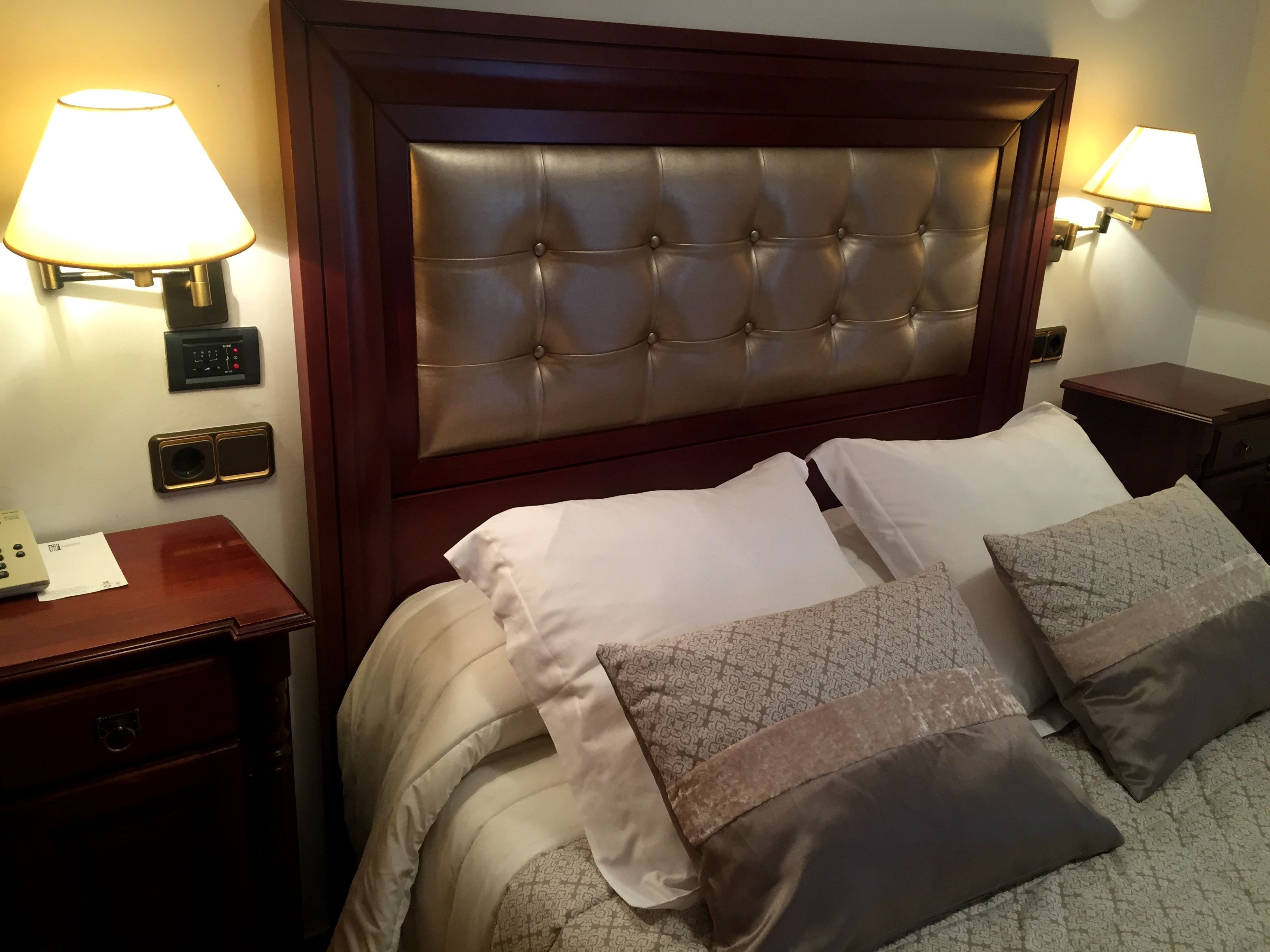 A hotel room with a tufted headboard, two bedside lamps, a bedside table with a phone and papers, and a bed with multiple pillows.