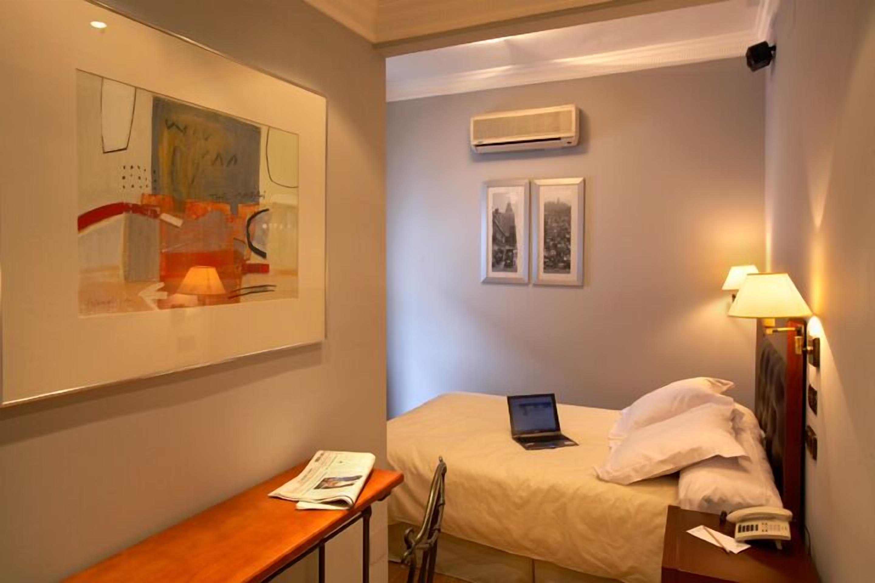 Habitación de hotel con cama, escritorio, ordenador portátil, lámpara y cuadros en la pared.