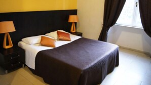 A neatly made bed with a dark bedspread and two pillows, flanked by two bedside tables with lamps, in a room with a yellow wall and a window with curtains.