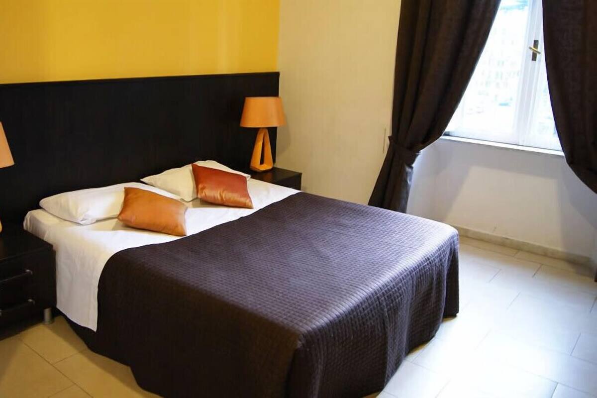 Ein ordentlich bezogenes Bett mit dunkler Bettdecke und zwei Kissen, flankiert von zwei Nachttischen mit Lampen, in einem Zimmer mit gelber Wand und einem Fenster mit Vorhängen.
