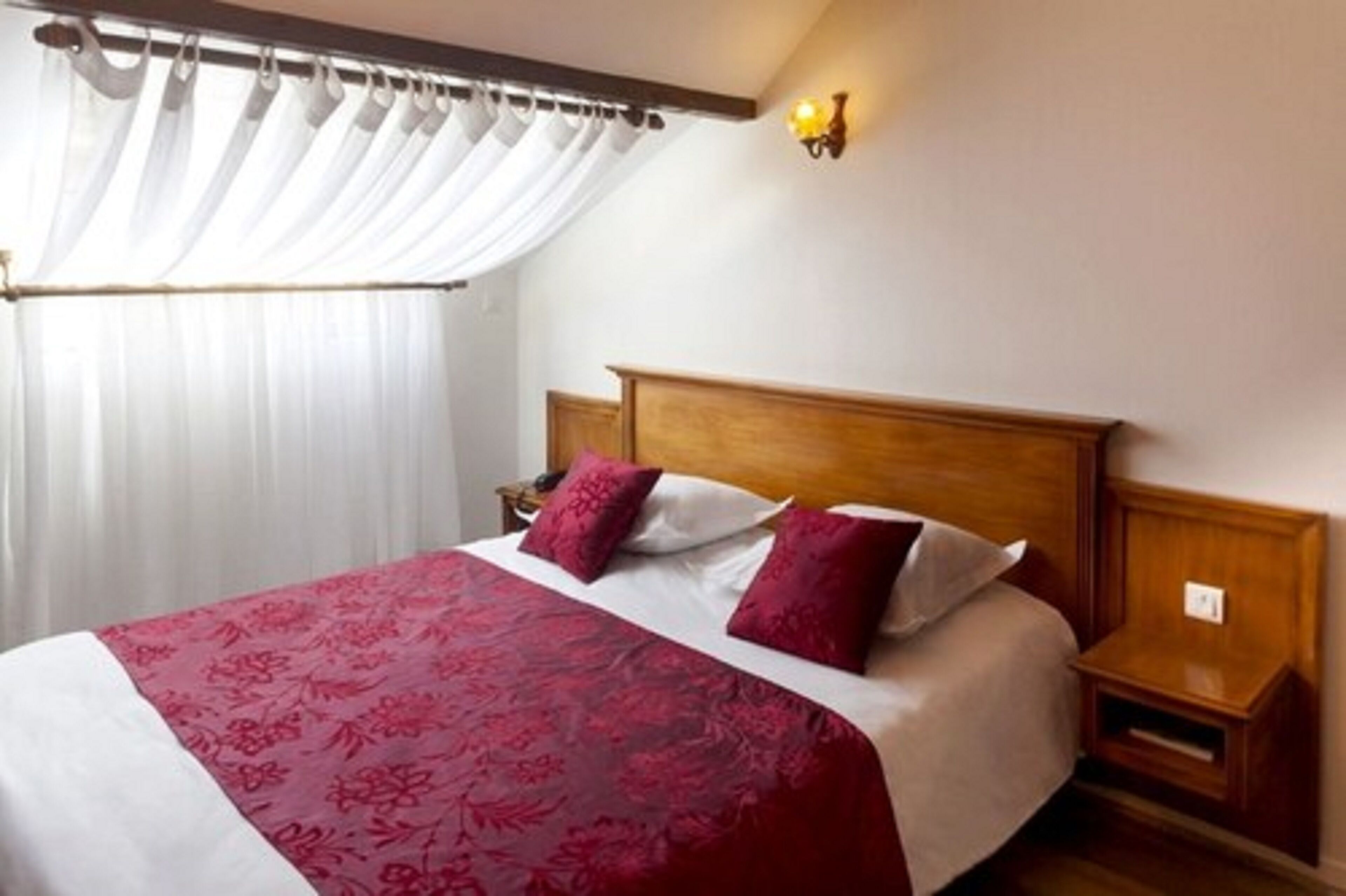 A hotel room with a large bed, white curtains, a wooden headboard, and a nightstand with a lamp.