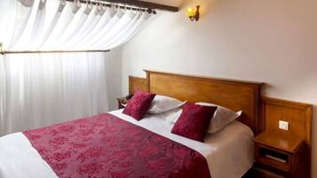 A hotel room with a large bed, white curtains, a wooden headboard, and a nightstand with a lamp.