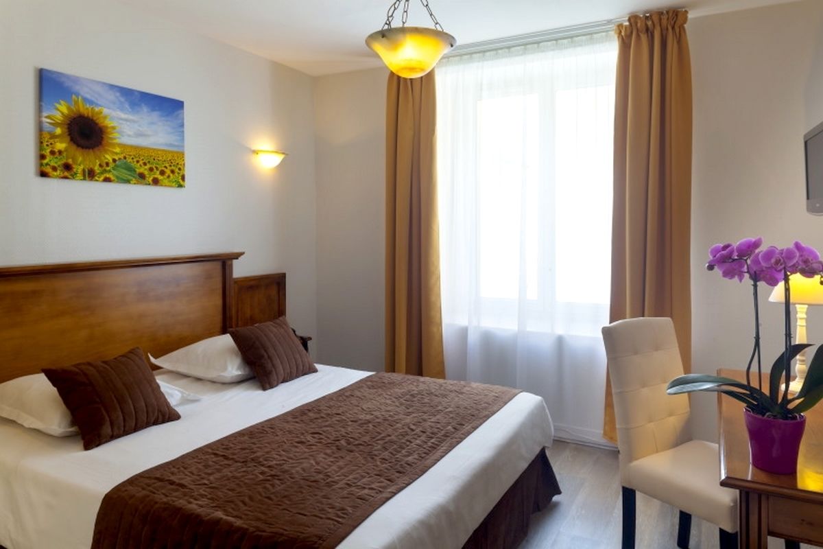 A hotel room with a bed, a chair, a table with a plant, a painting of a sunflower field, and a window with curtains.