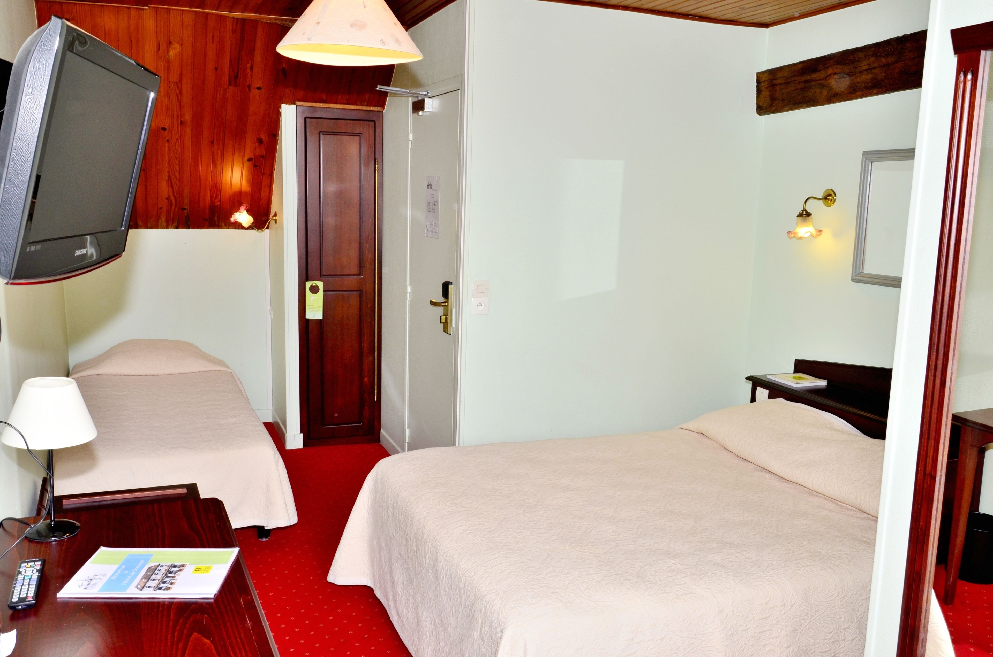 A hotel room with two beds, a TV mounted on the wall, a desk with a lamp, and a red carpet.