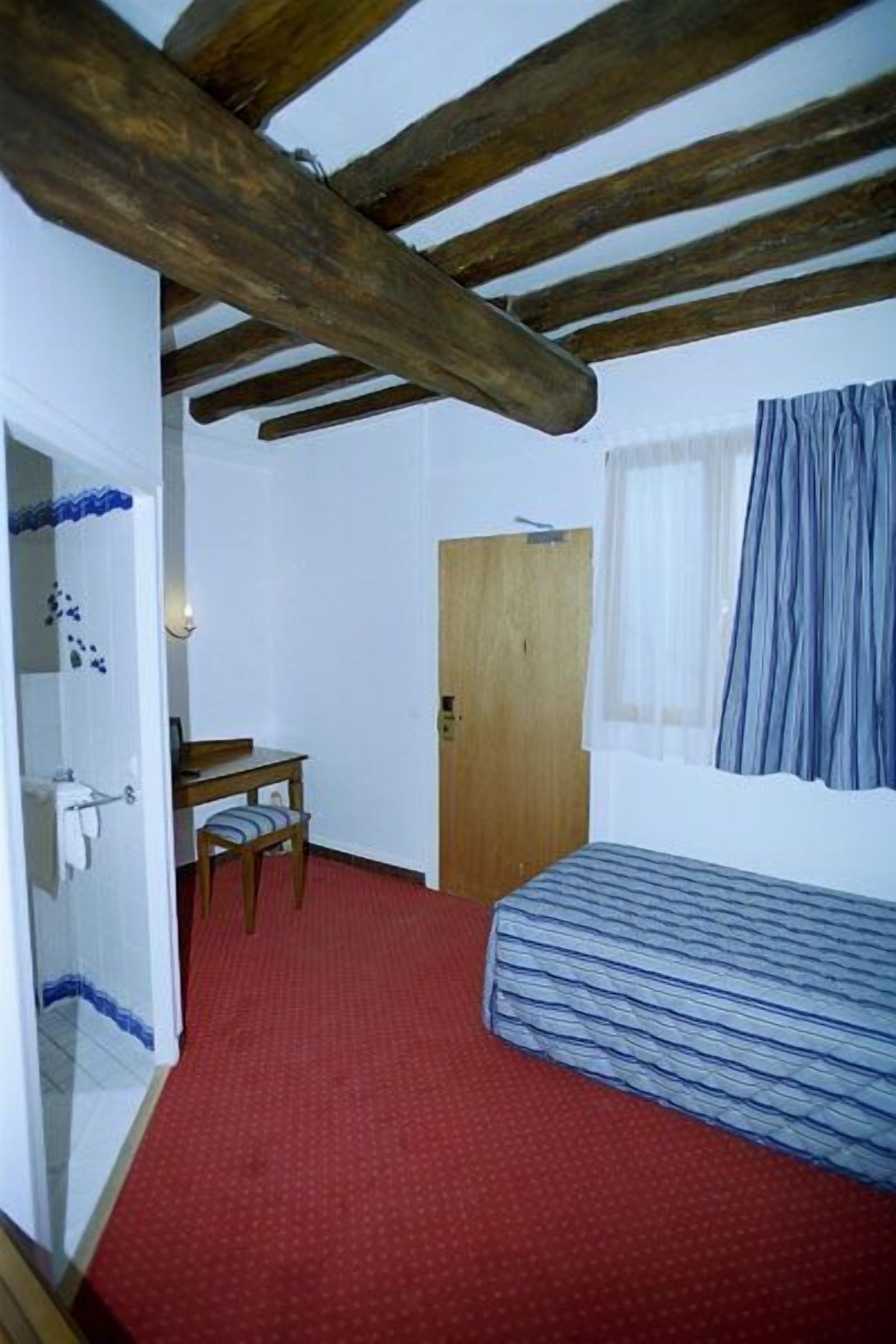 Quarto com teto de madeira, cama com cobertura listrada, uma mesinha com cadeira, porta de madeira e janela com cortinas.