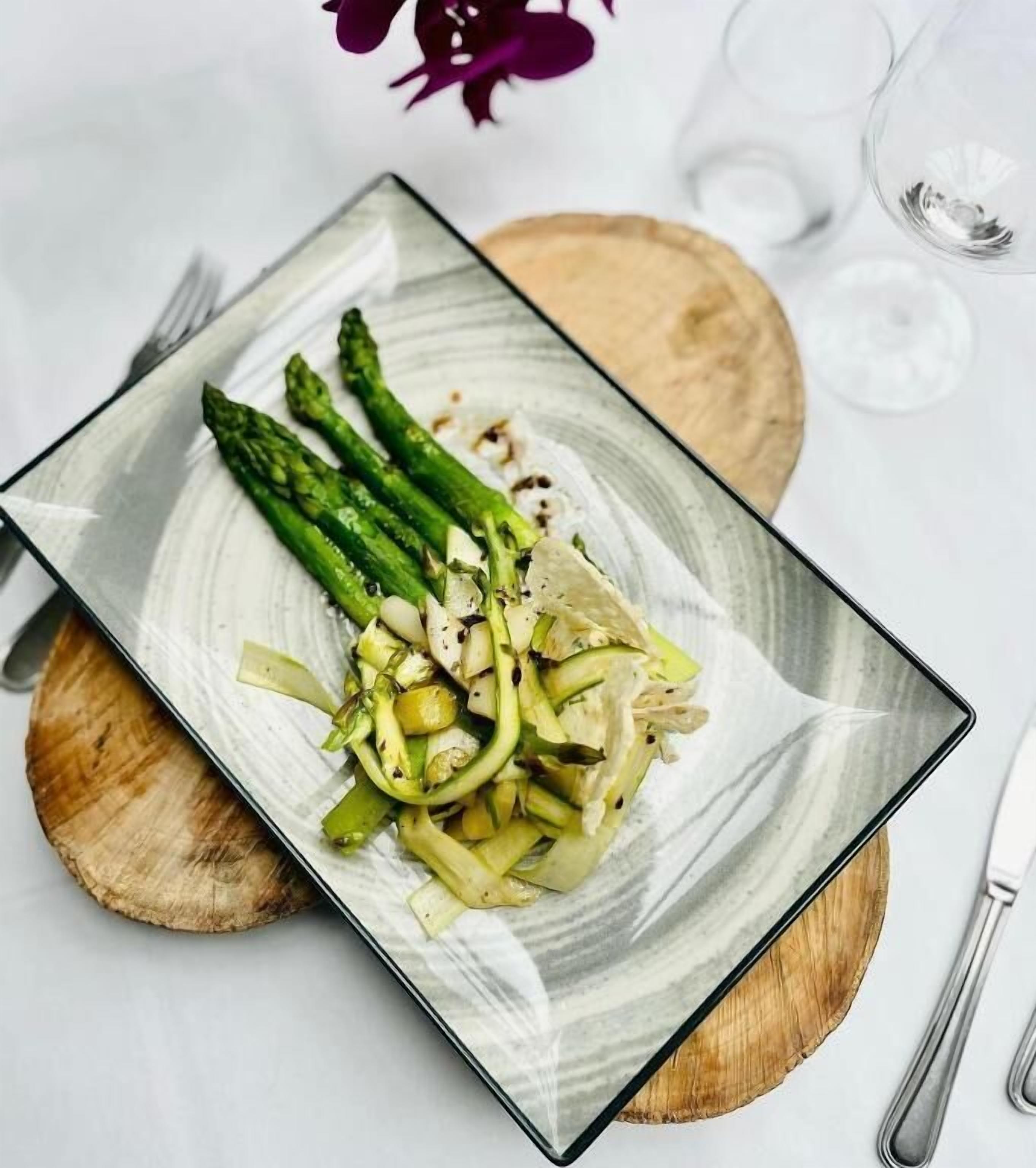 Ein Teller Essen mit Spargel und einem grünen Gemüse, möglicherweise Zucchini, auf einem rechteckigen Teller mit einem Holzuntersetzer darunter.