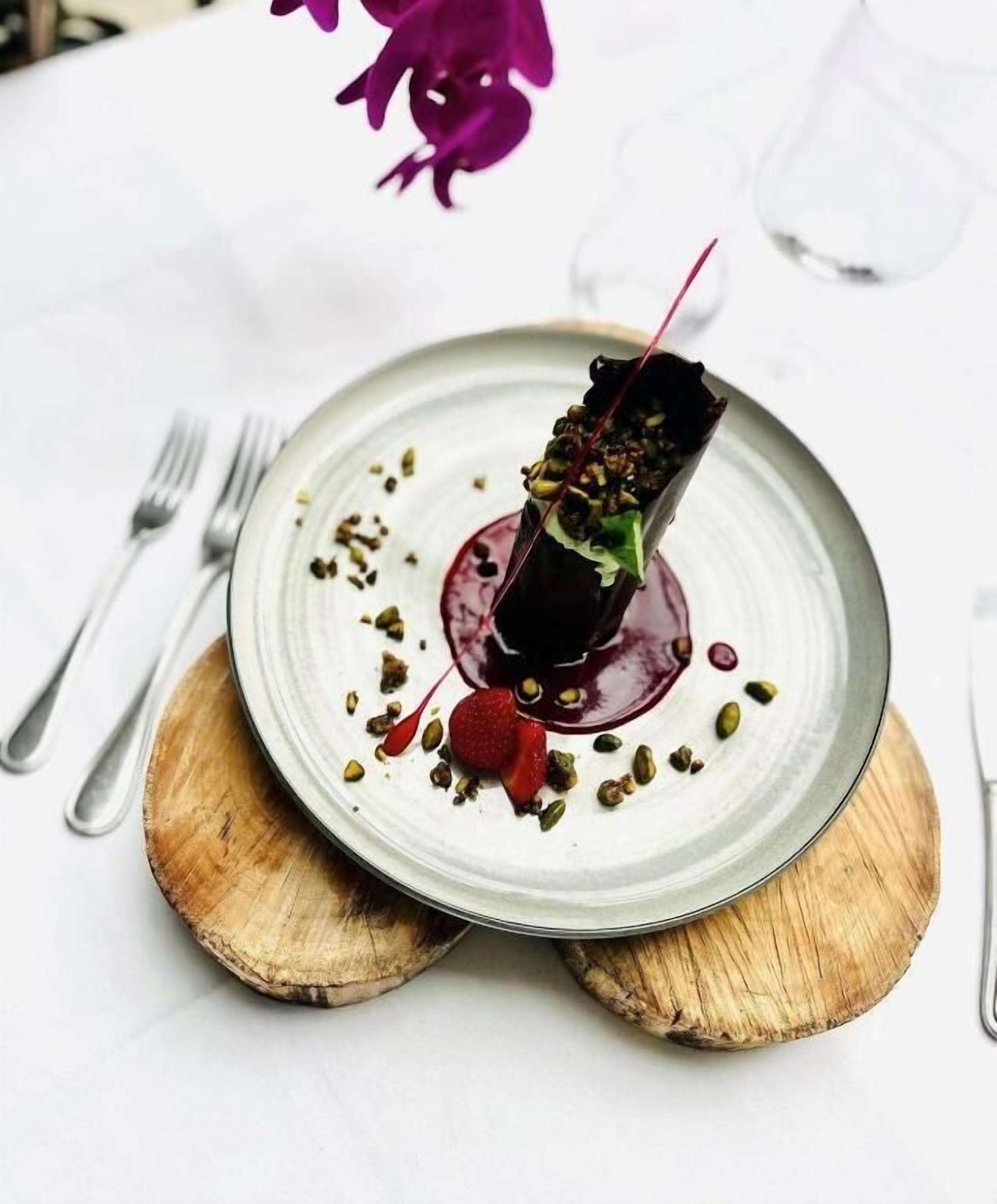 A plate with a dark-colored food item, garnished with green seeds and a red berry, on a white plate with a wooden base.