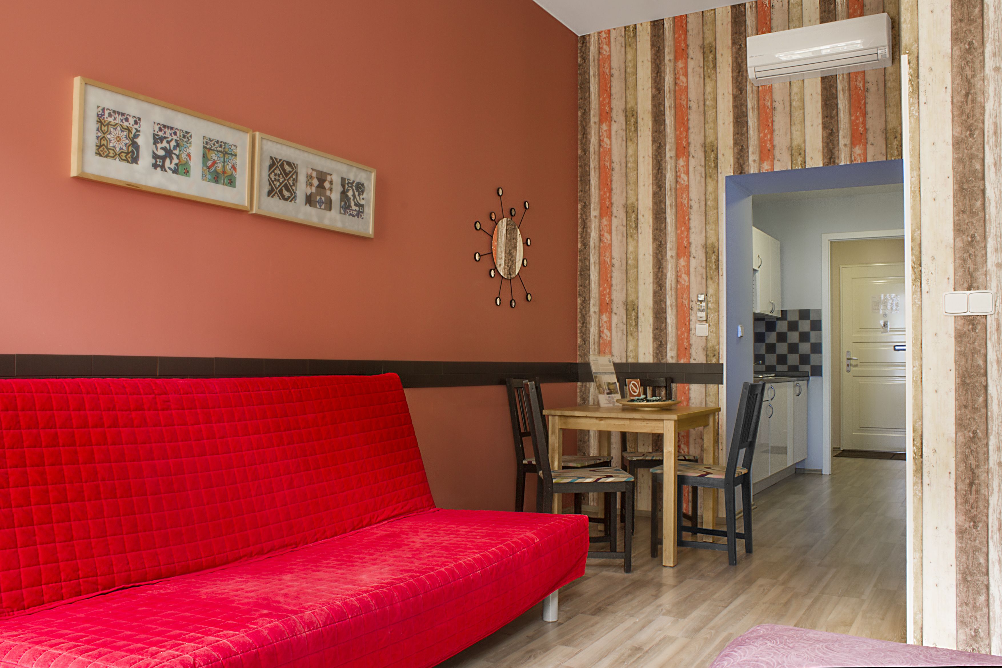 Un sofá rojo en una habitación con una mesa de comedor y sillas, un reloj en la pared, y una cocina visible a través de un vano abierto.