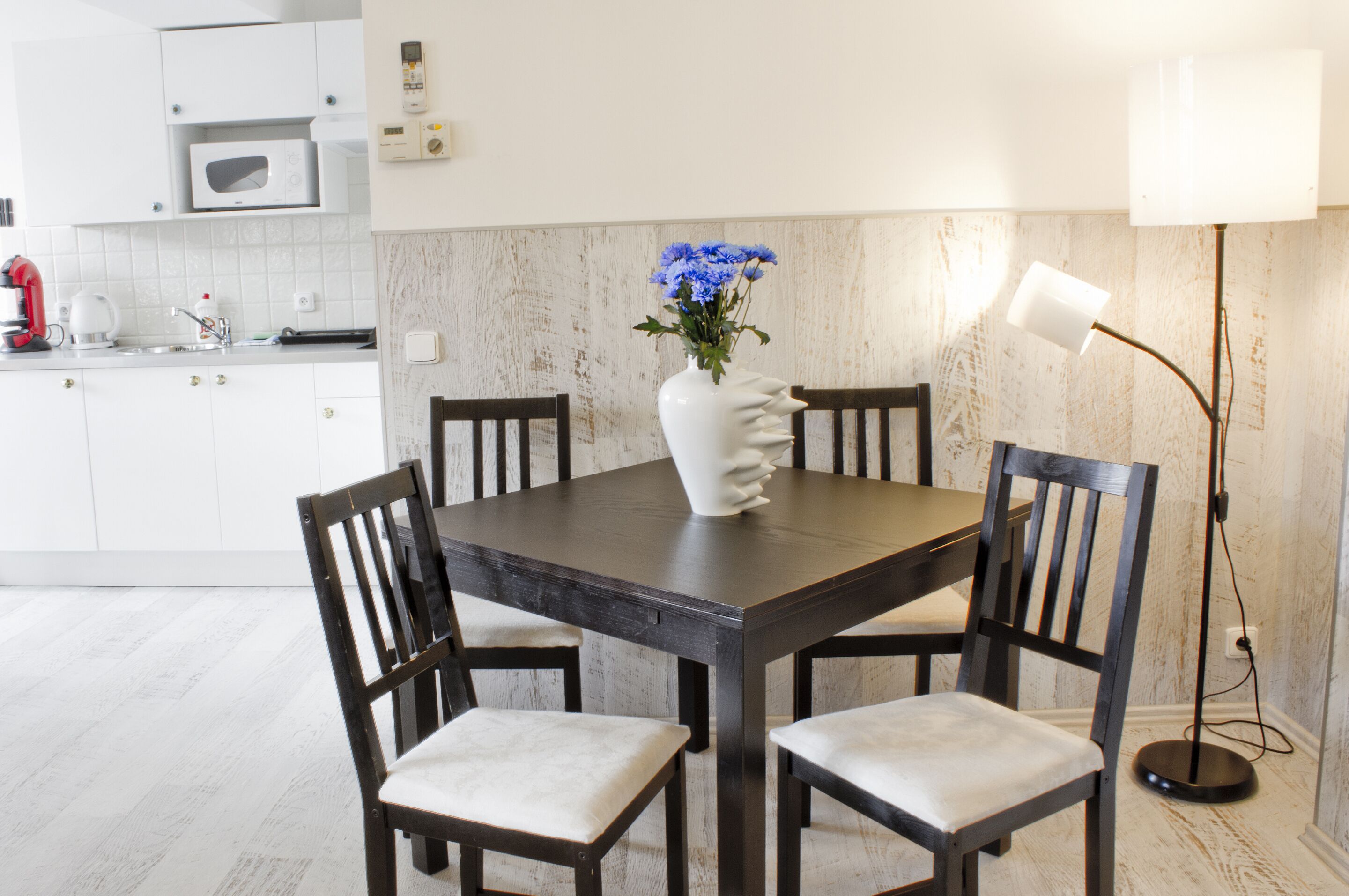 A dining table with four chairs, a white vase with flowers, a microwave, and a floor lamp.