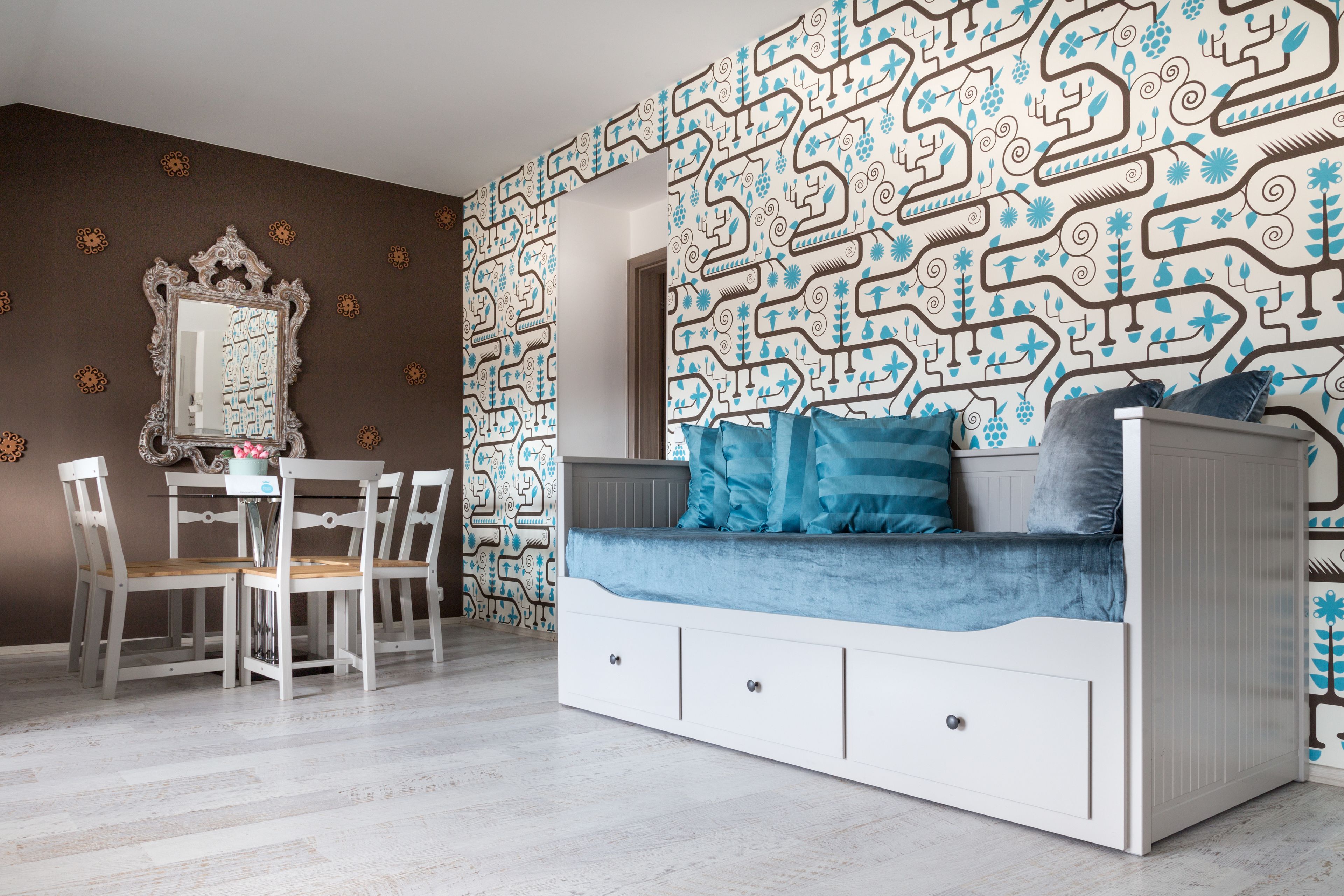 Um quarto com parede estampada, um sofá branco com almofadas azuis, uma mesa de jantar com cadeiras e um espelho na parede.
