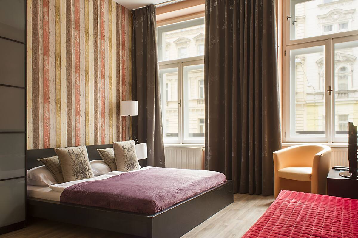 A hotel room with a bed, a chair, a window with curtains, and a view of buildings outside.