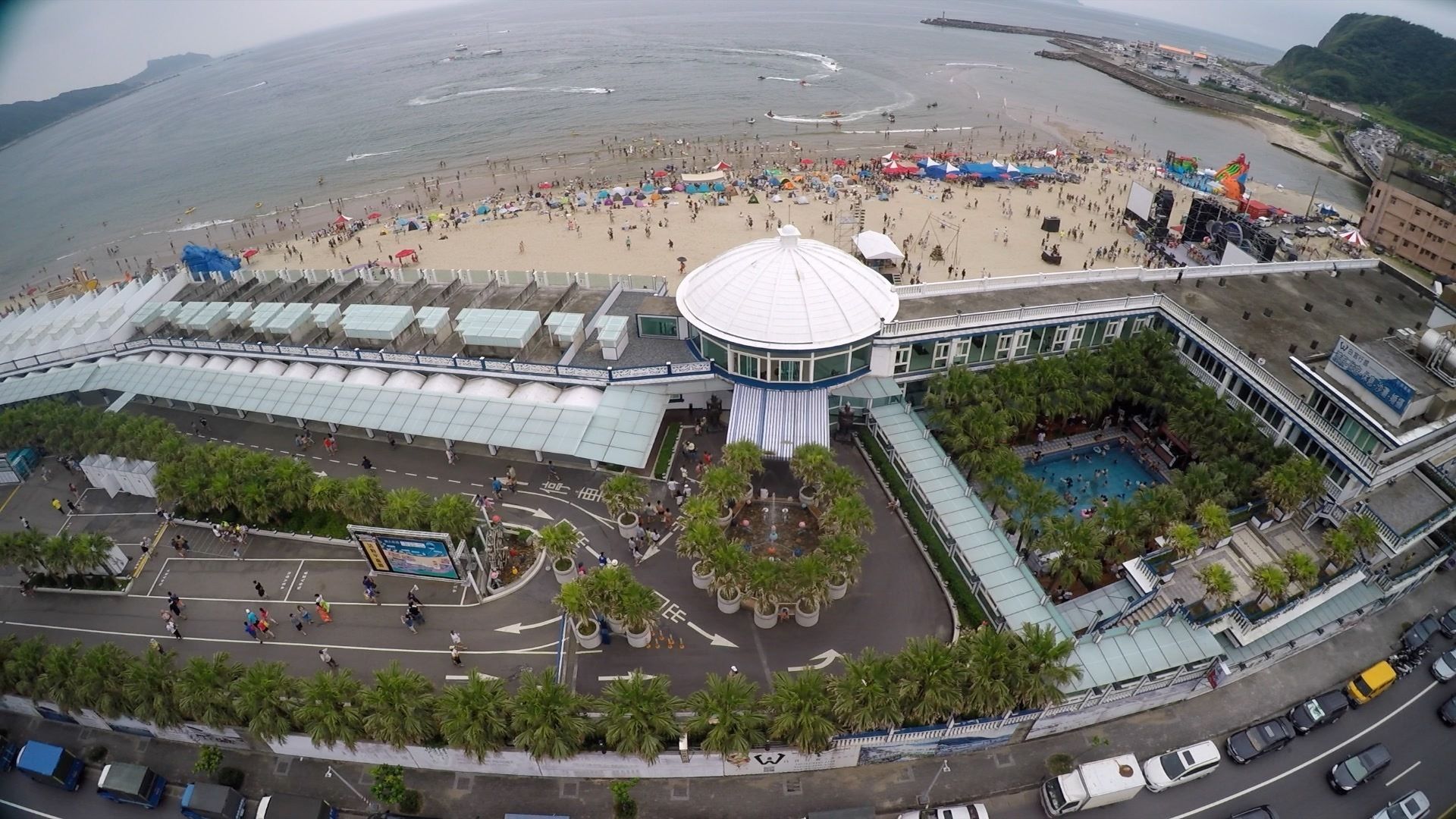 A large hotel with a central dome structure, a swimming pool, and a beach area with umbrellas.