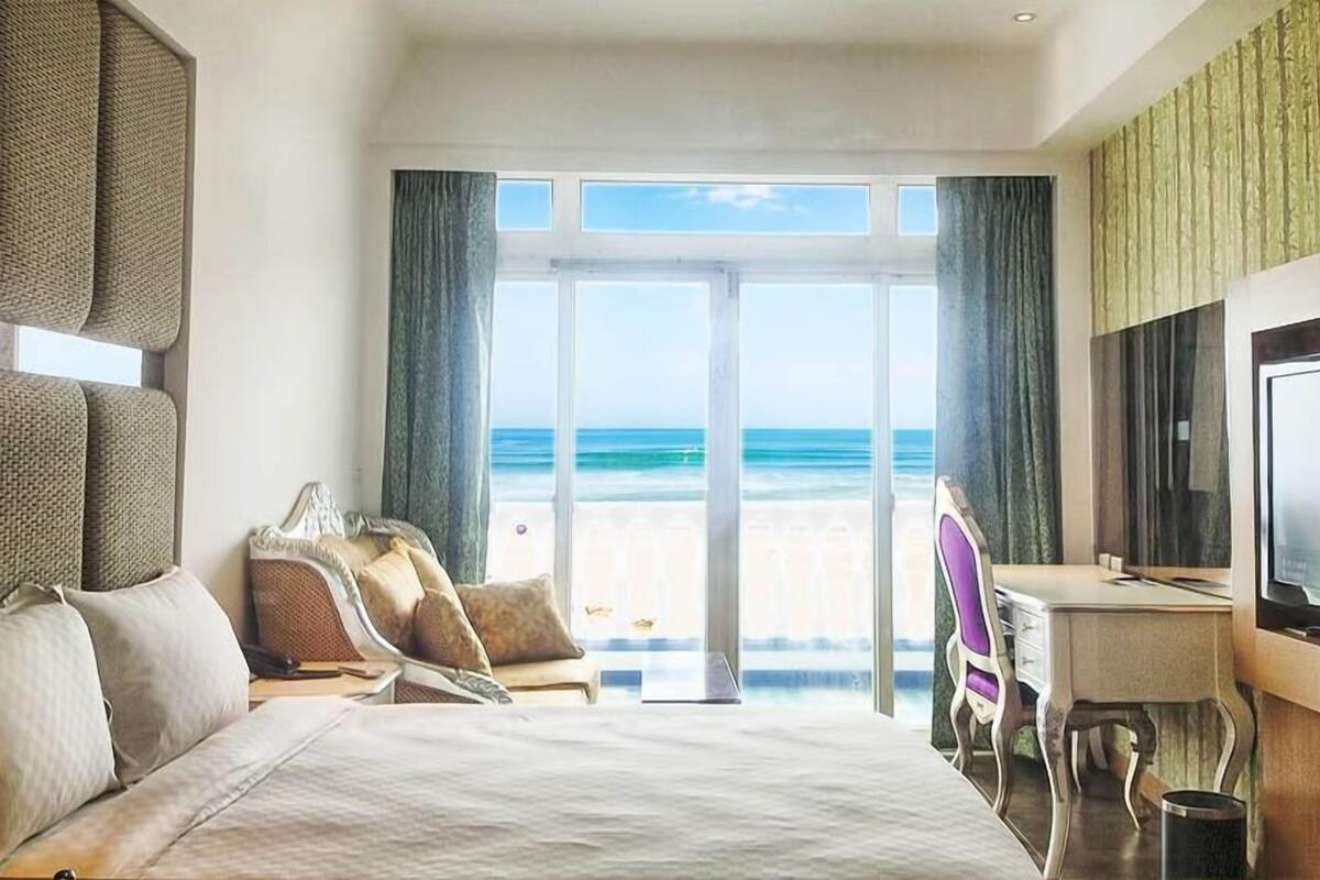 A hotel room with a bed, a desk, and a television. The room has a view of the beach and ocean.