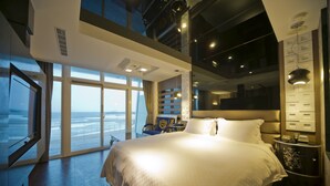 Habitación de hotel moderna con una cama grande, una mesita de noche, una silla y vista al océano a través de amplias ventanas.
