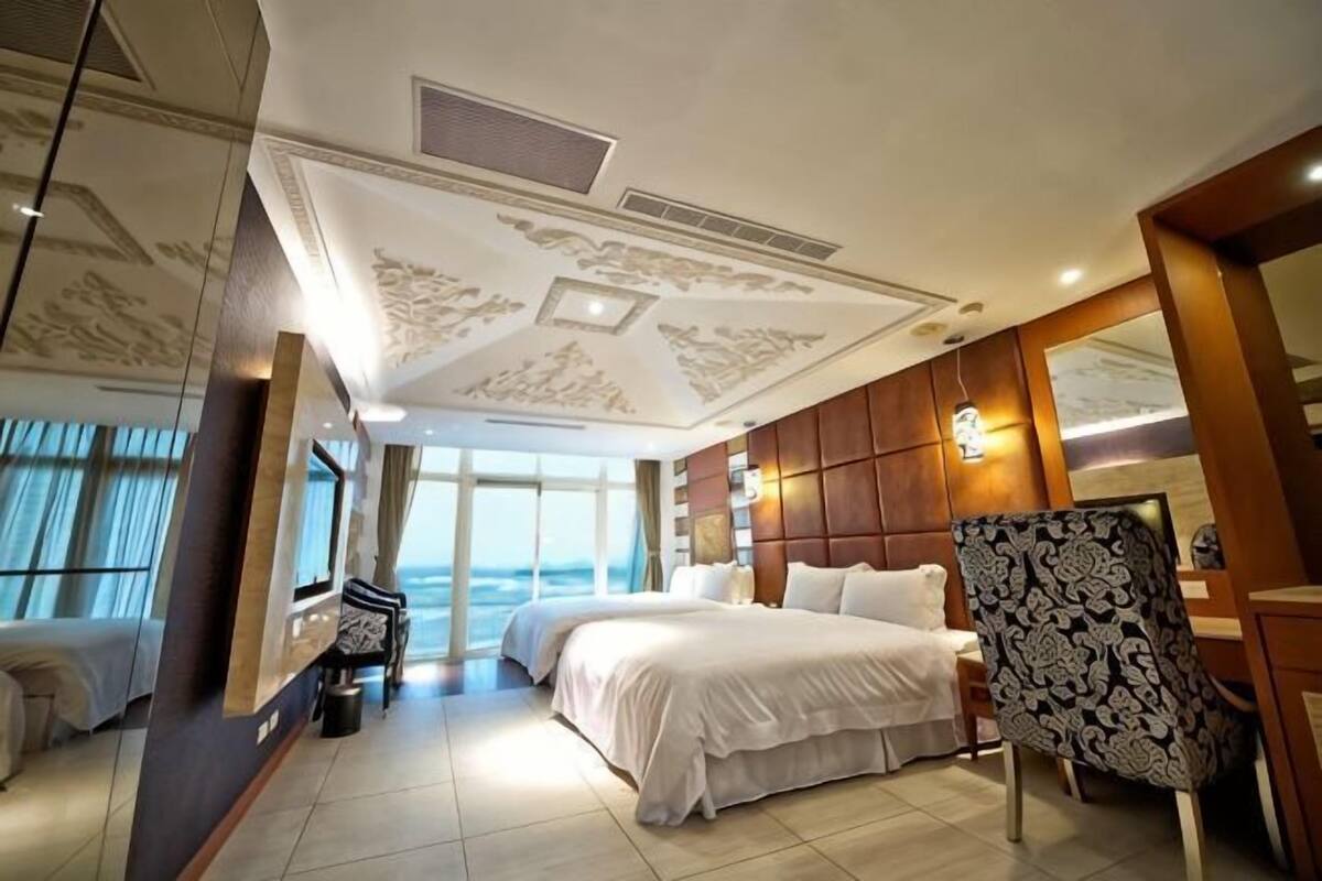 A hotel room with a large bed, a sitting area with a chair, a desk, and a view of the sea.