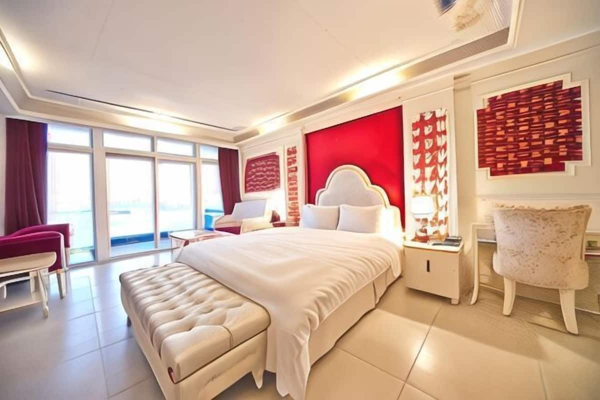 A modern hotel room with a large bed, a sitting area with a red chair, and a view through the window.