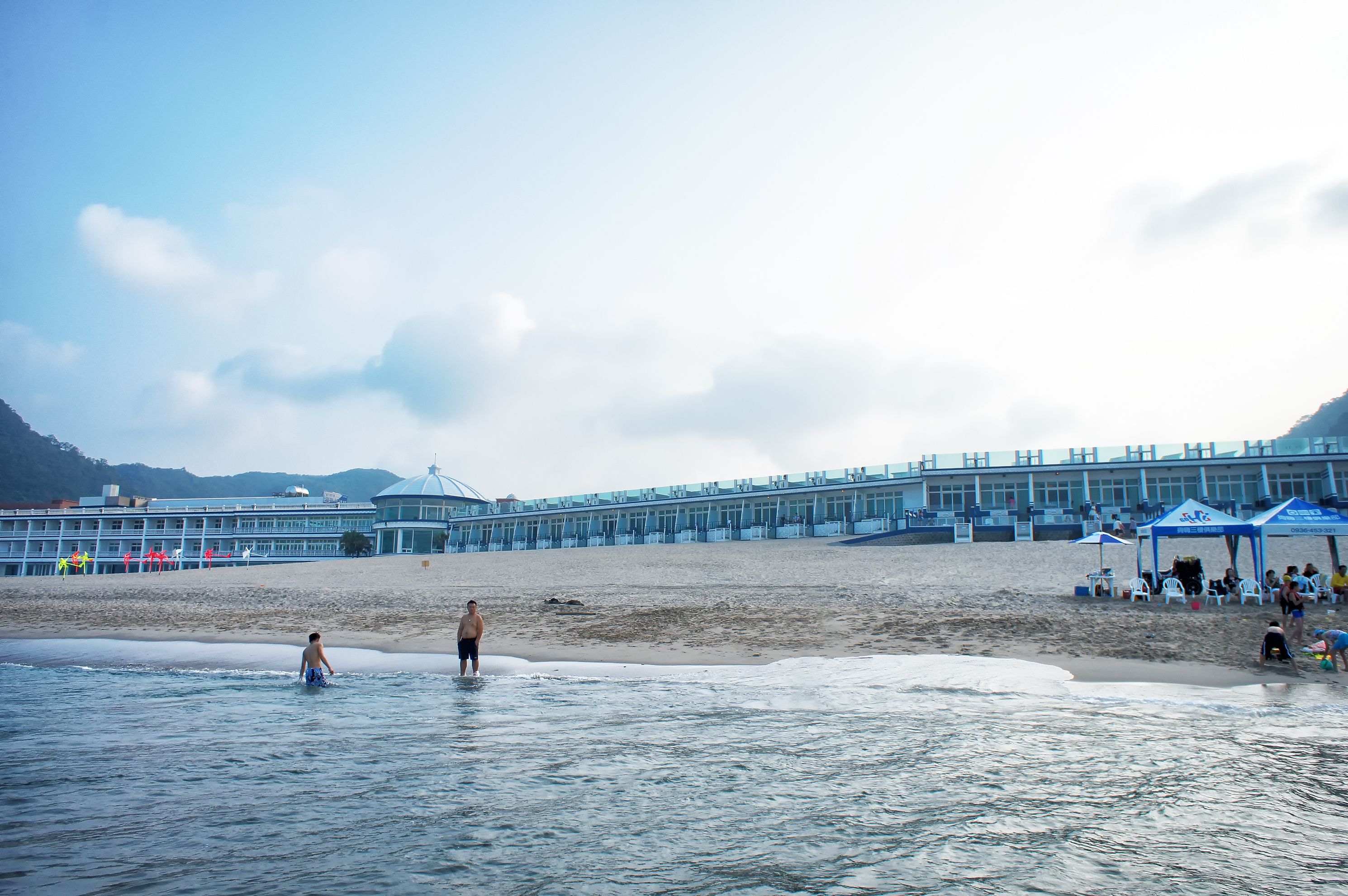 Una playa con un edificio grande al fondo, una orilla arenosa y algunas personas en el agua.