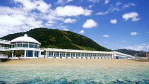 Un hotel frente a la playa con un edificio circular, un largo pasillo con ventanas y montañas al fondo.
