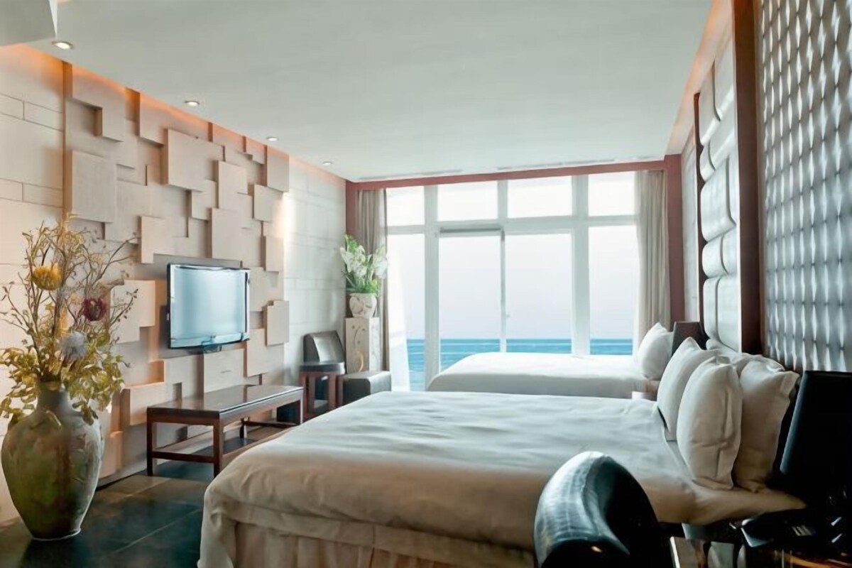 A hotel room with a bed, a TV, a desk, and a view of the sea.