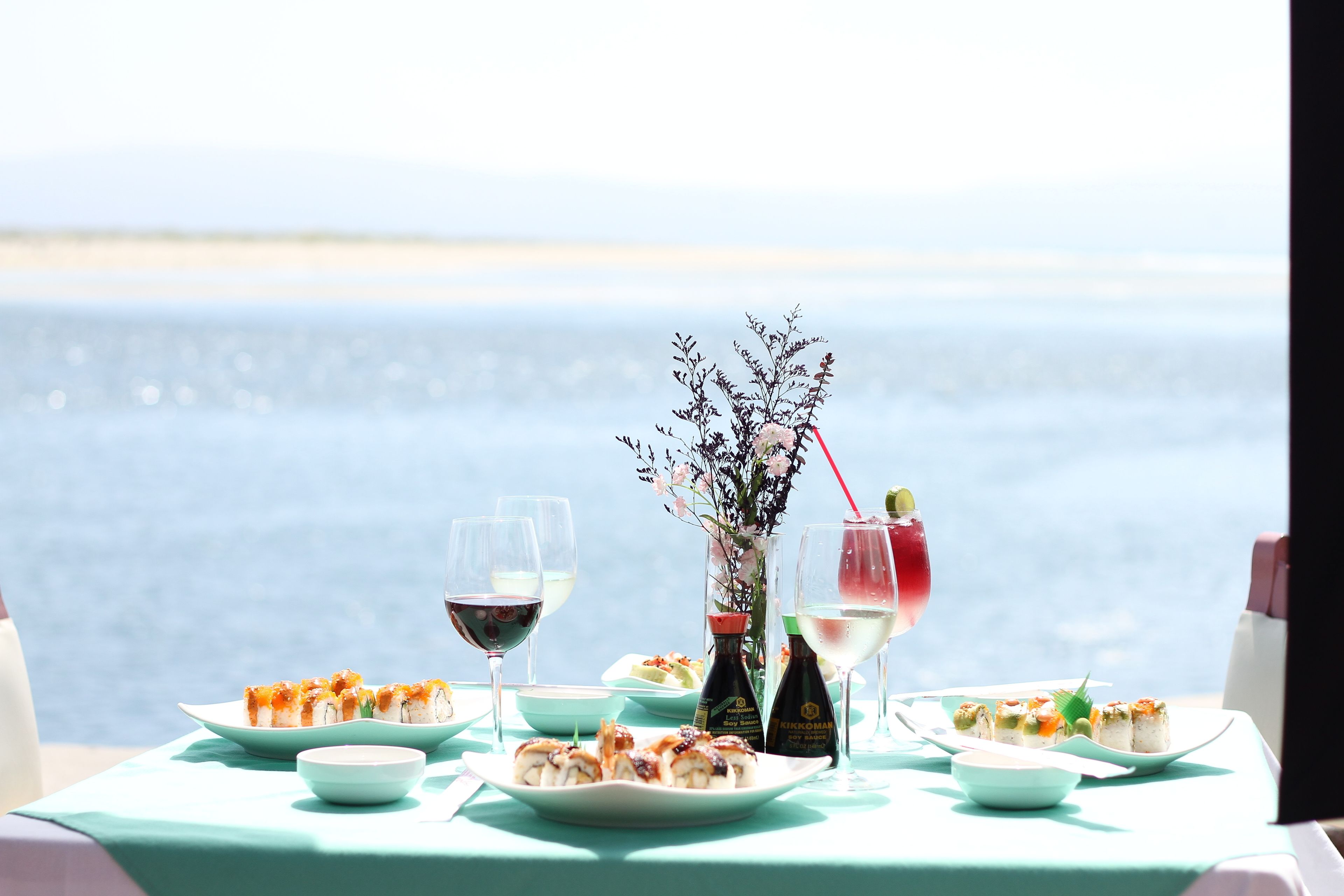 A table set with sushi, wine glasses, and a vase with flowers, overlooking a body of water.