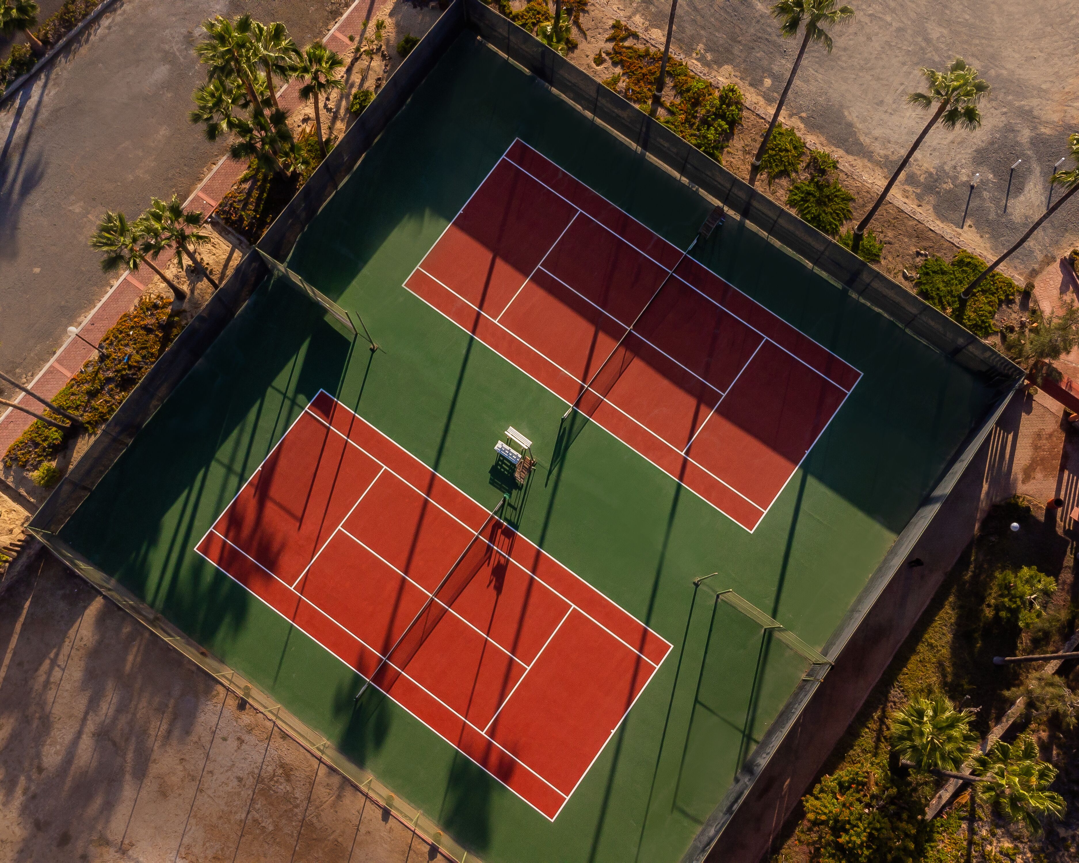 Luftaufnahme eines Tennisplatzes mit umliegenden Palmen und einem gepflasterten Weg.