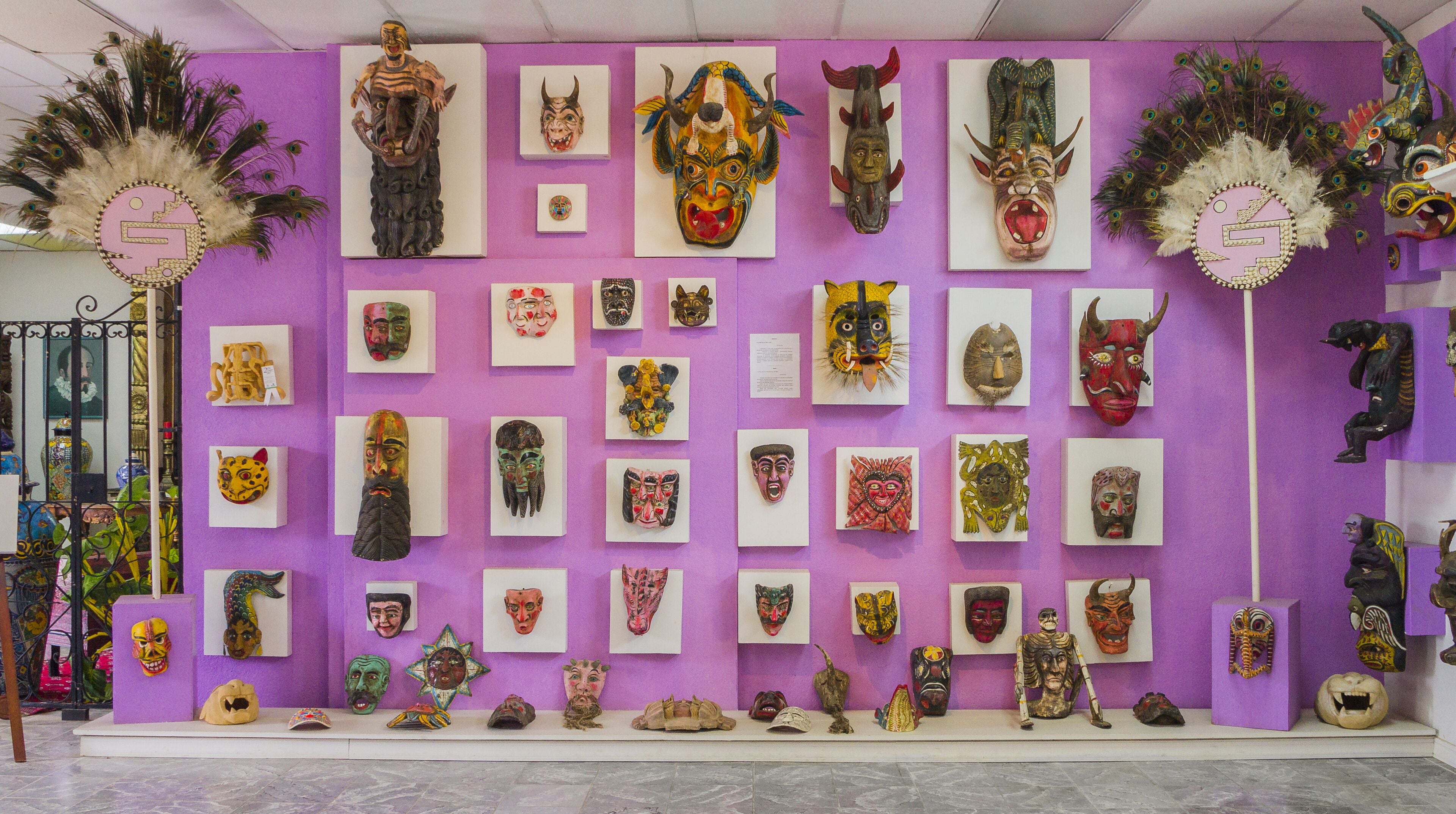 Een muur met een verzameling traditionele maskers en artefact, gerangschikt in een raster op een paarse achtergrond.