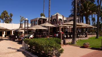 An outdoor dining area with multiple tables and umbrellas, surrounded by palm trees and a well-maintained lawn.