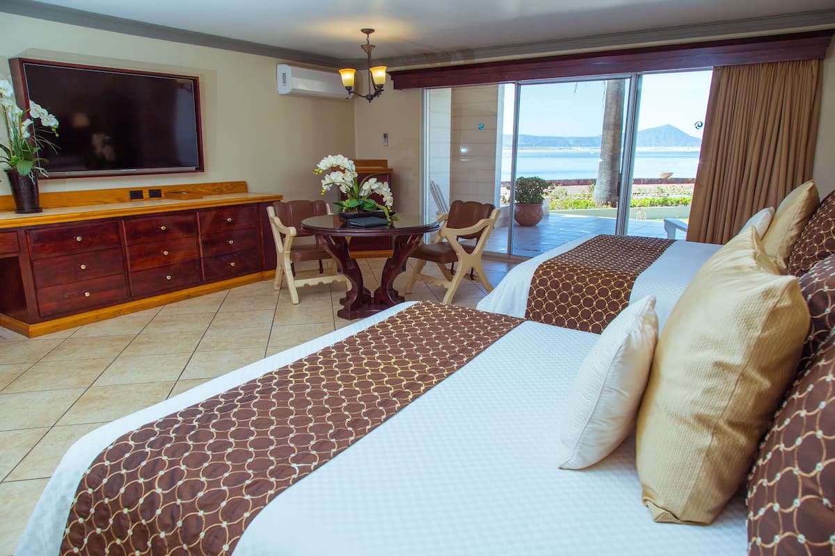 A hotel room with a large bed, a TV, a dining table with chairs, and a view of the sea.