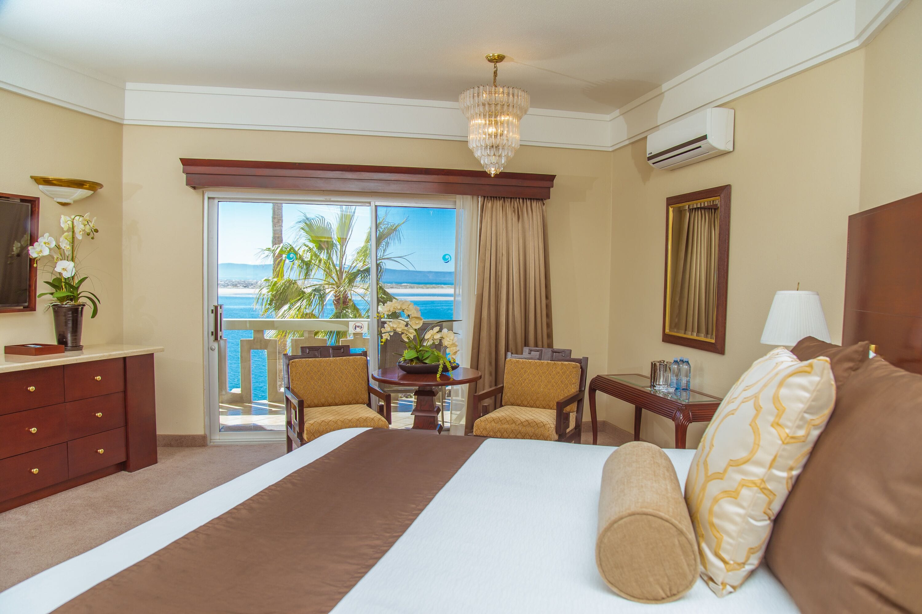 A hotel room with a large bed, a desk, chairs, a view of the ocean, and a balcony.