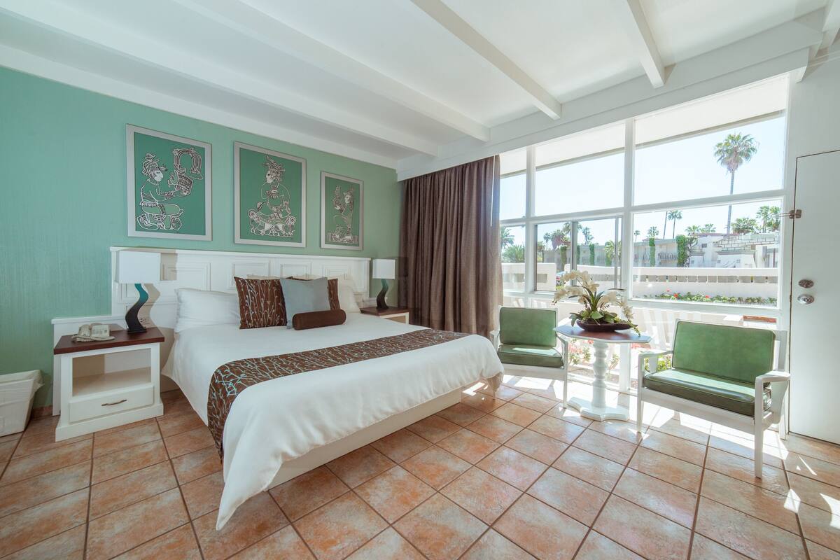 A hotel room with a large bed, a small table with a plant, a green armchair, and a view of palm trees through large windows.