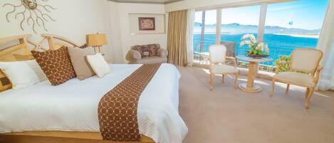 A hotel room with a large bed, a sofa, a dining table, and a view of the sea.