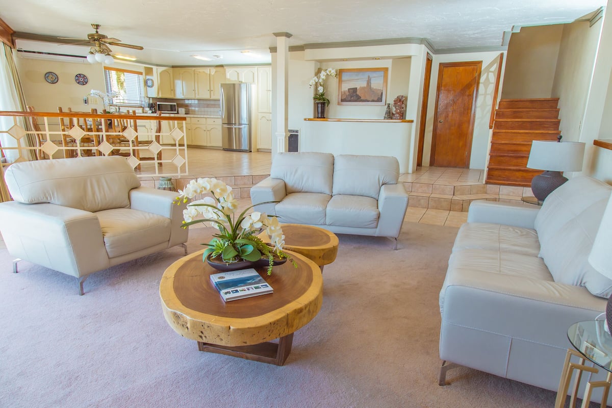 A spacious living area with a beige sofa, a round wooden coffee table with a magazine, and a potted orchid. A kitchen is visible in the background with a refrigerator and cabinets.