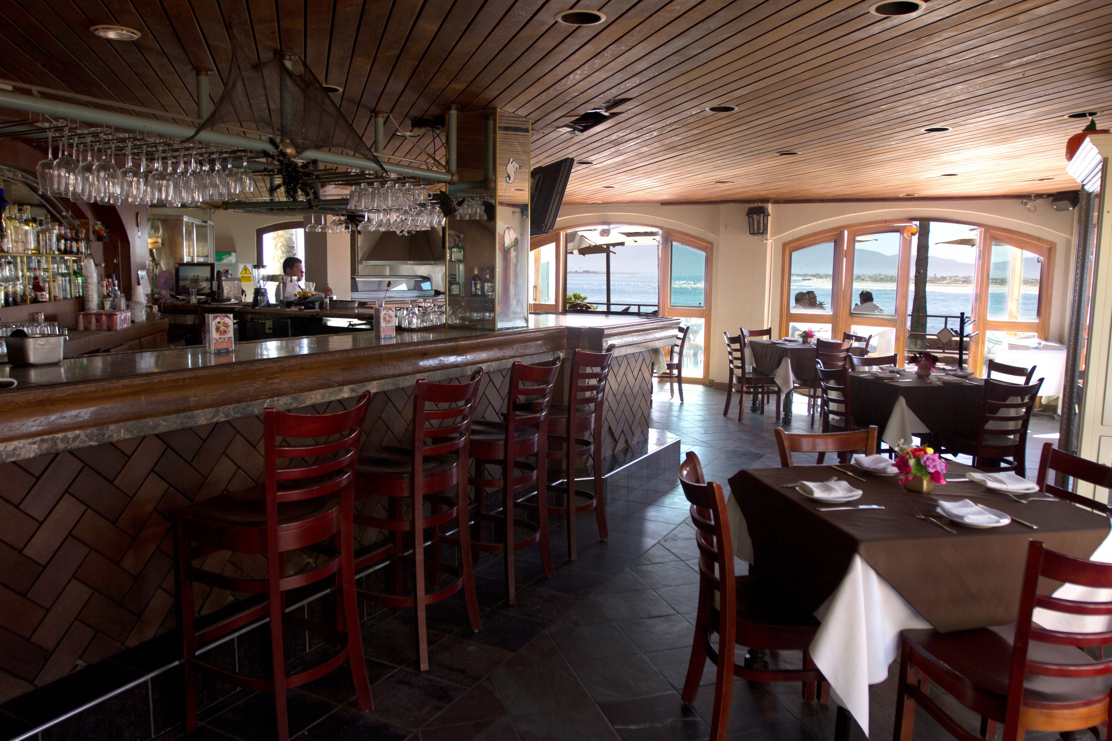 Ein Restaurant mit Barbereich, verschiedenen Sitzmöglichkeiten, darunter hohe Stühle und Tische, und großen Fenstern mit Meerblick.